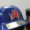 A glossy blue baseball helmet featuring a UV DTF sticker transfer with a bold red "A" outlined in white. The sticker seamlessly adheres to the curved surface, showcasing its durability and vibrant finish. In the background, a black tumbler with colorful lettering is visible.
