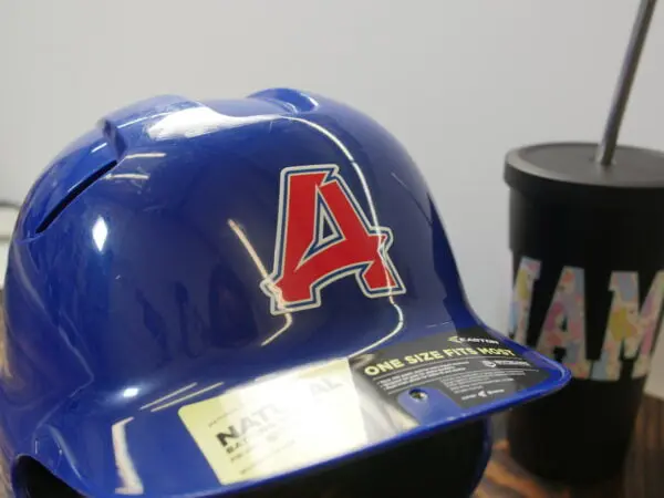 A glossy blue baseball helmet featuring a UV DTF sticker transfer with a bold red "A" outlined in white. The sticker seamlessly adheres to the curved surface, showcasing its durability and vibrant finish. In the background, a black tumbler with colorful lettering is visible.
