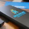 A black portable charger featuring a UV DTF sticker transfer with a bold blue "A" logo, a gold crown, and the text "Arizona Ink Custom Print Solutions." The raised, glossy finish of the sticker highlights the vibrant colors and durability of UV DTF printing. A USB flash drive and blue background elements are partially visible on a wooden surface.