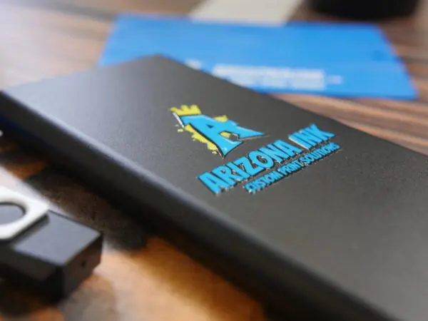 A black portable charger featuring a UV DTF sticker transfer with a bold blue "A" logo, a gold crown, and the text "Arizona Ink Custom Print Solutions." The raised, glossy finish of the sticker highlights the vibrant colors and durability of UV DTF printing. A USB flash drive and blue background elements are partially visible on a wooden surface.