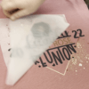 A DTF transfer being peeled off a pink t-shirt after heat pressing. The design features bold black text with gold accents and decorative splashes, showcasing the vibrant colors and sharp details of Direct-to-Film printing on fabric.