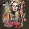 A DTF-printed graphic t-shirt featuring a vibrant, stylized portrait of Marilyn Monroe. The design includes bold red lips, detailed shading, and a graffiti-style background with splashes of yellow, blue, and pink. The word "Marilyn" is scripted in bright red at the bottom, showcasing the high-quality color vibrancy and detail achieved with DTF printing on fabric.