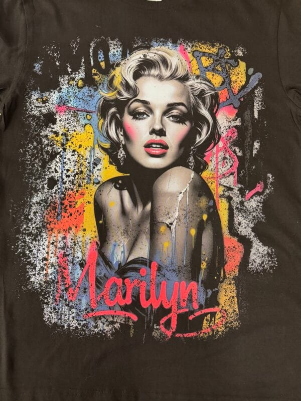 A DTF-printed graphic t-shirt featuring a vibrant, stylized portrait of Marilyn Monroe. The design includes bold red lips, detailed shading, and a graffiti-style background with splashes of yellow, blue, and pink. The word "Marilyn" is scripted in bright red at the bottom, showcasing the high-quality color vibrancy and detail achieved with DTF printing on fabric.