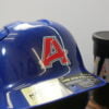 A glossy blue baseball helmet featuring a UV DTF sticker transfer with a bold red "A" outlined in white. The sticker seamlessly adheres to the curved surface, showcasing its durability and vibrant finish. In the background, a black tumbler with colorful lettering is visible.