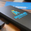 A black portable charger featuring a UV DTF sticker transfer with a bold blue "A" logo, a gold crown, and the text "Arizona Ink Custom Print Solutions." The raised, glossy finish of the sticker highlights the vibrant colors and durability of UV DTF printing. A USB flash drive and blue background elements are partially visible on a wooden surface.