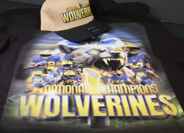 A DTF-printed t-shirt featuring a Wolverines National Champions design with a fierce, growling wolverine, a group of football players in blue and yellow uniforms, and a championship trophy. The bold yellow and blue text pops against the dark background, showcasing the sharp details and vibrant colors of DTF printing. A matching beige and black cap with the same Wolverines National Champions design is placed on top.